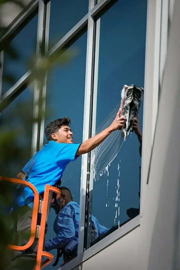 Window cleaners, one spraying soapy water, working on large glass windows of a building. Blue sky reflected.