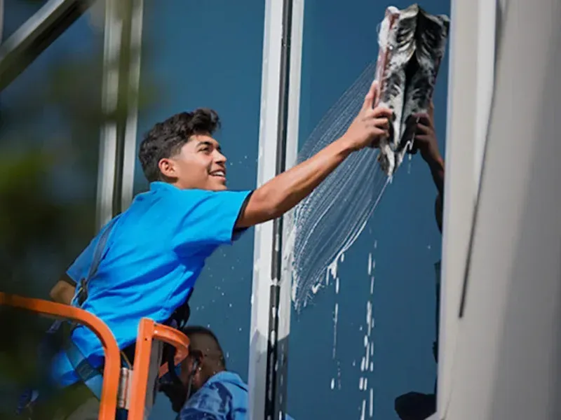 Window cleaner on lift, washing exterior window with a squeegee; blue shirt, sunny day.