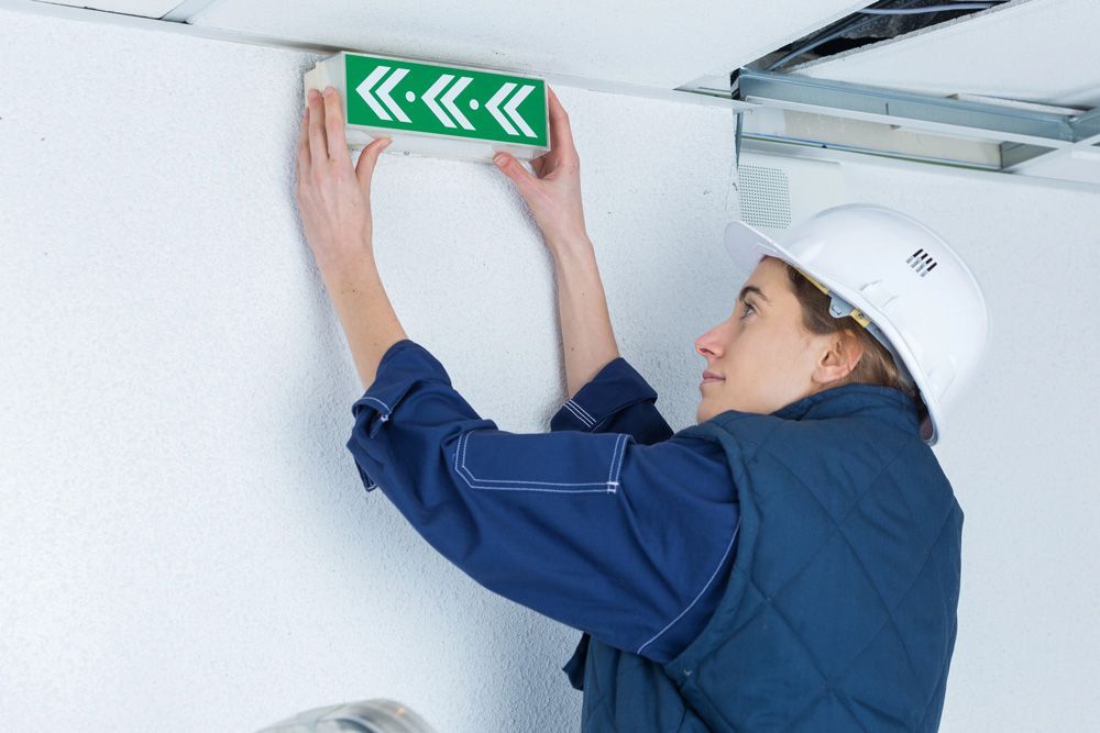 Woman in hard hat installs an exit sign on a white wall.