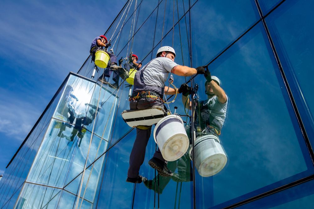 Window cleaners in safety harnesses suspended by ropes on the exterior of a glass skyscraper against a blue sky.