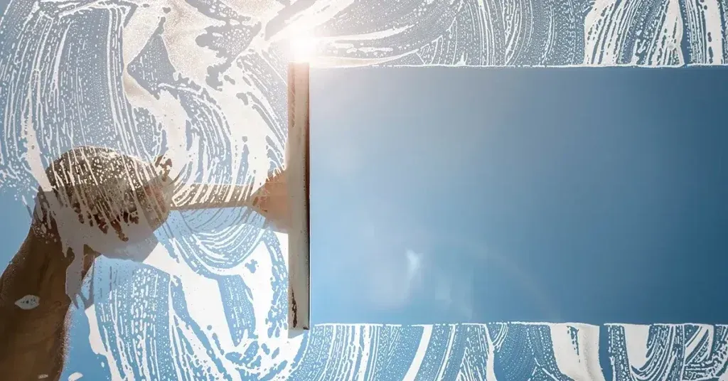 Person cleaning a window with soapy water, sky visible through the clean pane.