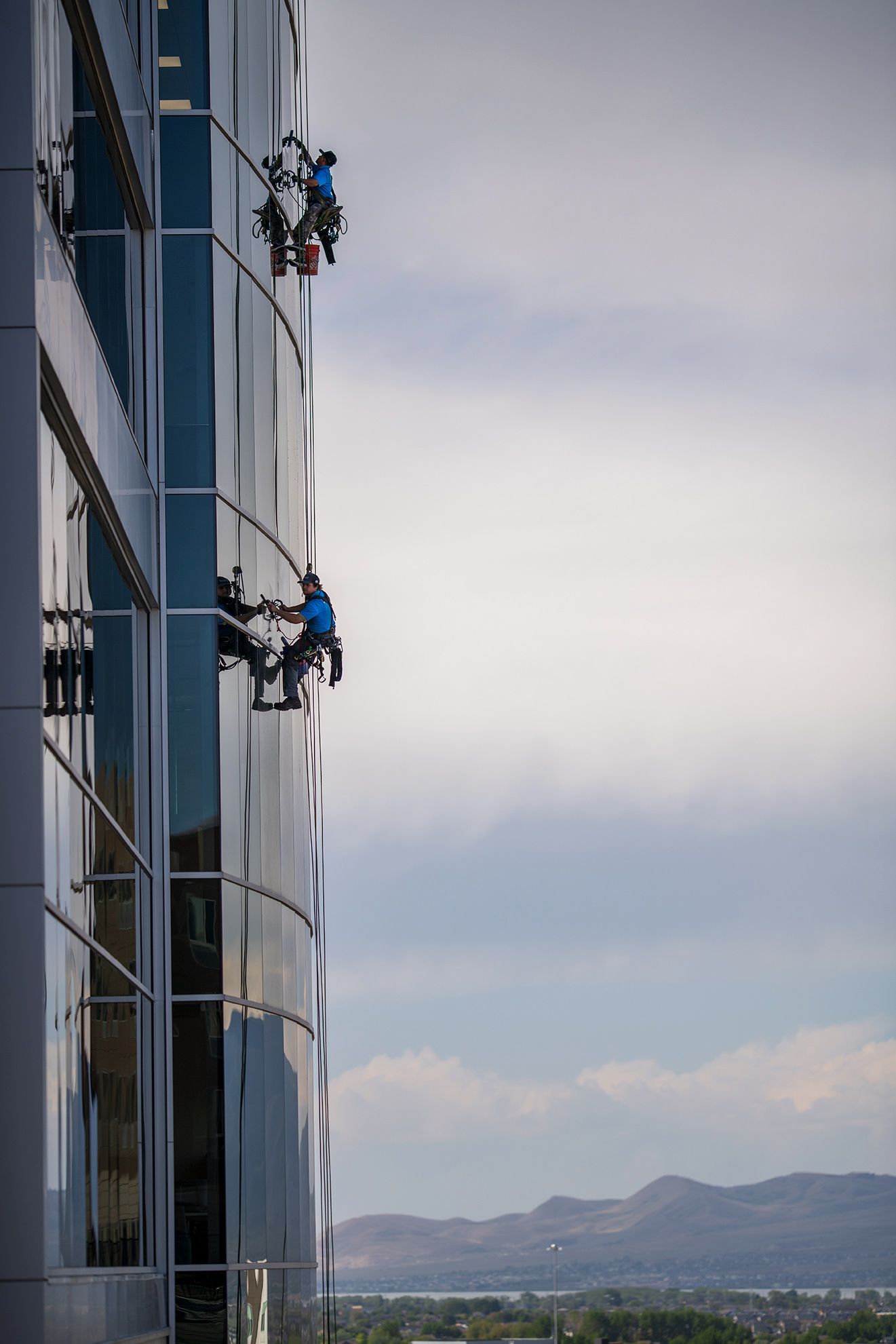 Two window cleaners in blue shirts suspended by ropes cleaning the glass exterior of a high-rise building.