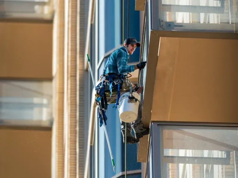 Window washer suspended on ropes cleaning an exterior window of a modern building.