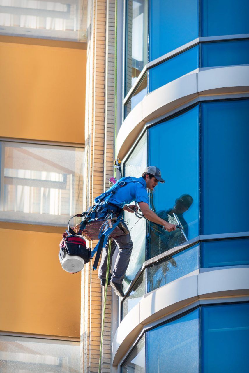 Window washer cleaning a tall, blue glass building with safety harness.