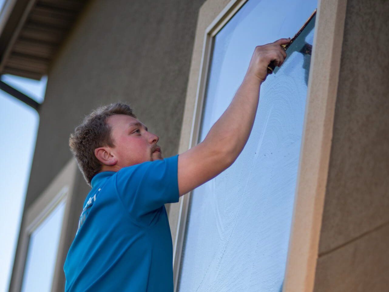 A person in a blue shirt uses a squeegee to clean a tall glass window on a building exterior.