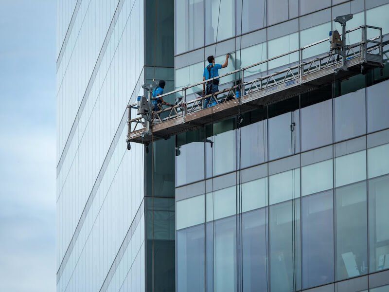 Three workers in blue shirts clean the glass windows of a high-rise office building from a suspended scaffold.