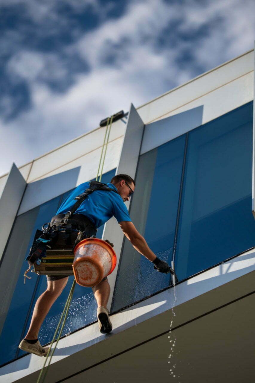 Window washer rappelling down a building, cleaning glass.  Blue shirt, harness, bucket, cloudy sky.