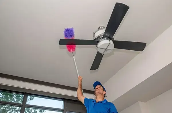 Person in blue shirt using a duster to clean a ceiling fan near a window.