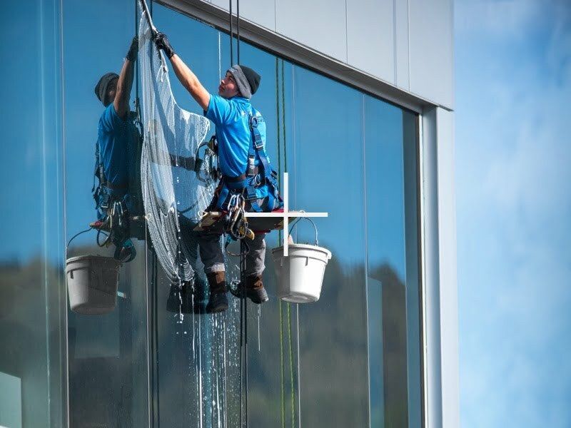 A worker in a blue shirt suspended by ropes cleans the large glass windows of a modern building with a squeegee.
