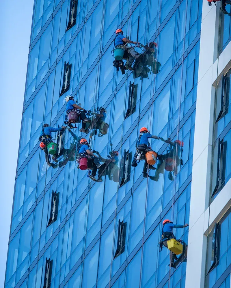 Window cleaners on ropes, washing the glass facade of a tall, blue-tinted skyscraper.