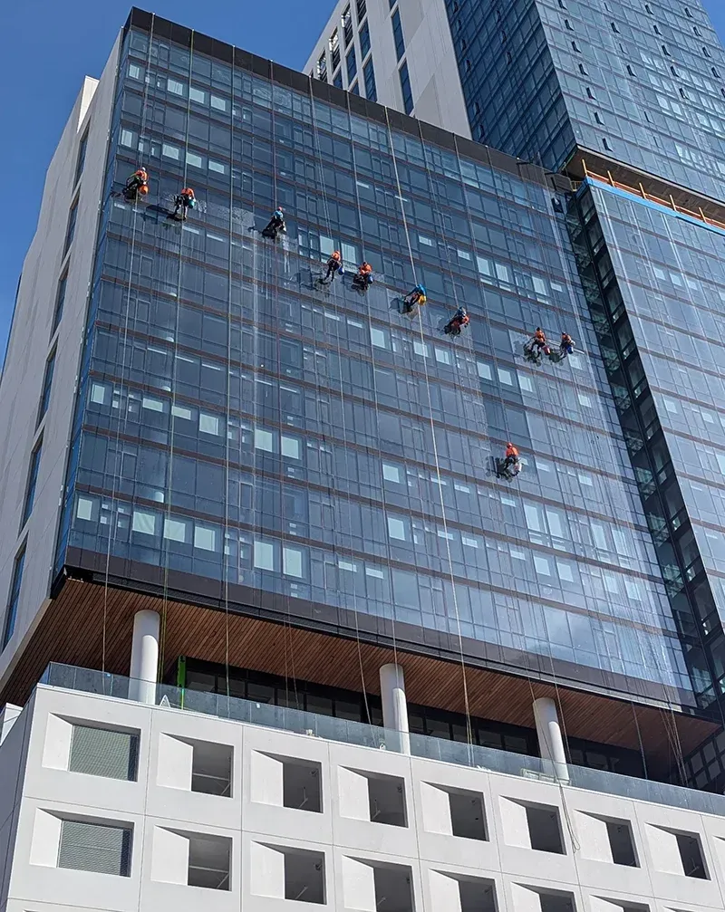 Workers cleaning windows of a high-rise building, suspended by ropes. Blue glass facade against a clear sky.