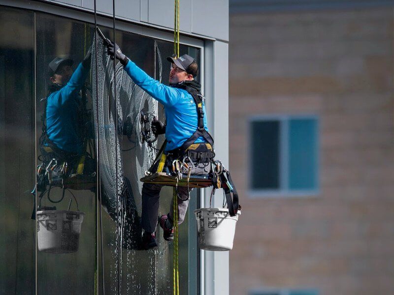 A professional window washer in a blue jacket suspended by ropes against a building, cleaning glass with a squeegee.