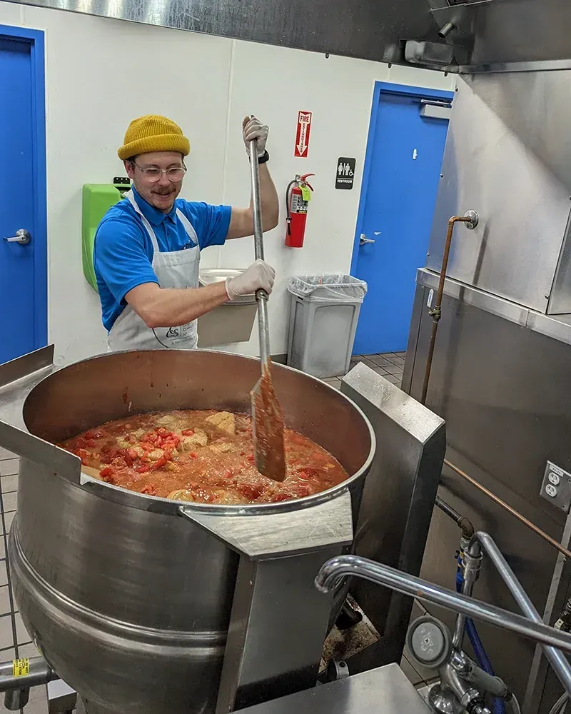 Chef stirs large pot of food in commercial kitchen. He wears a blue shirt, apron, and yellow hat.