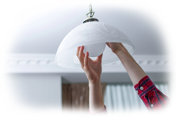 A person in a red plaid shirt reaches inside a white frosted glass ceiling lamp shade to install a light bulb.