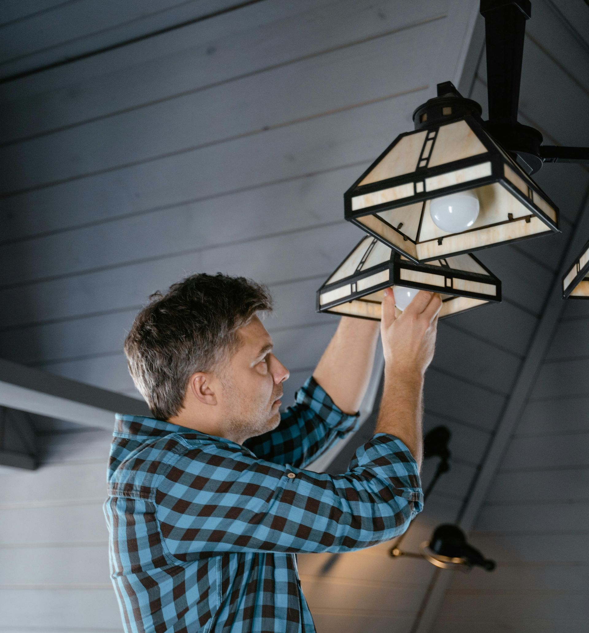 Man in plaid shirt replacing a lightbulb in a chandelier, indoors.