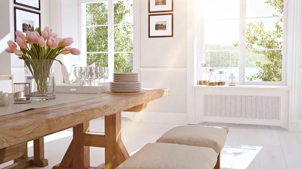 Bright dining room with a wooden table, bench seating, and a vase of pink tulips. Sunlight streams in through windows.