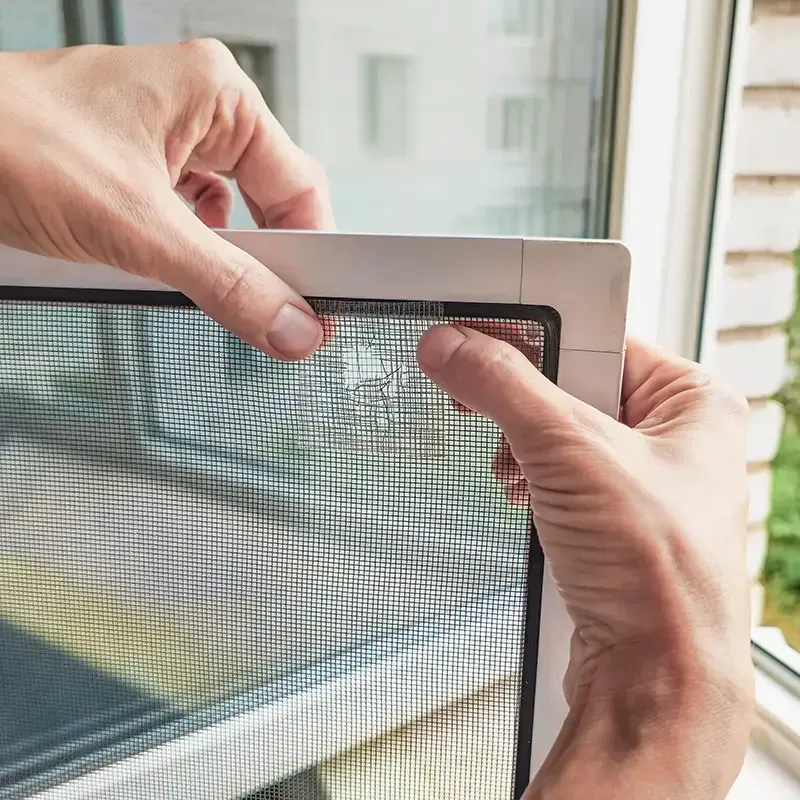 Hands installing a gray mesh window screen in a window frame