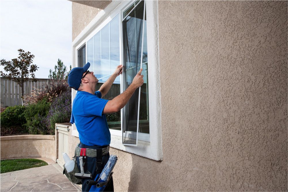 Man in blue shirt and cap installing window screen on a stucco house exterior.