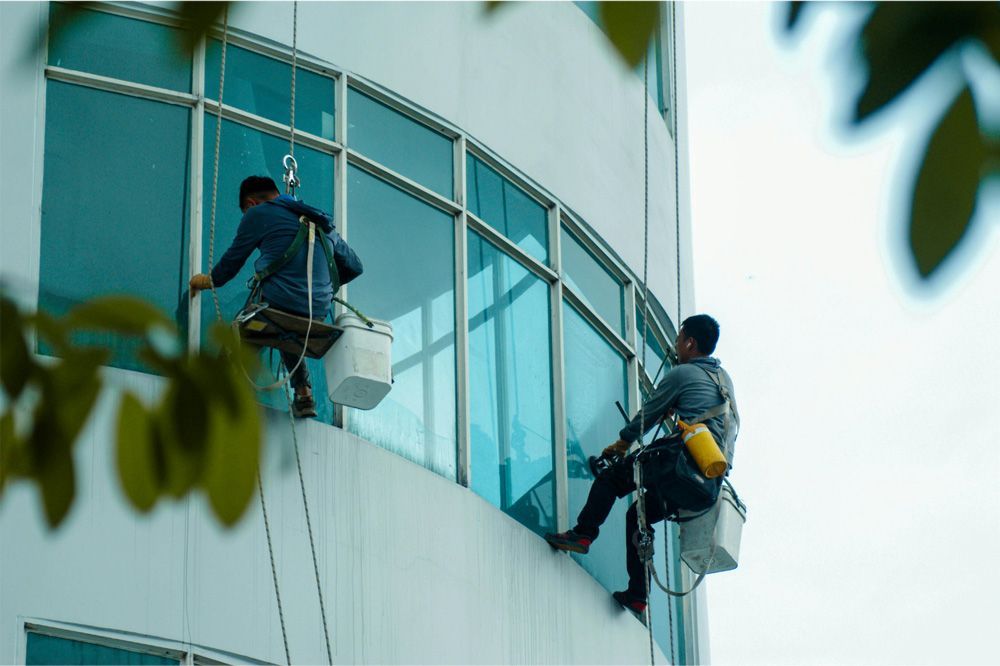 Two window washers on a curved building, suspended by ropes, cleaning glass.