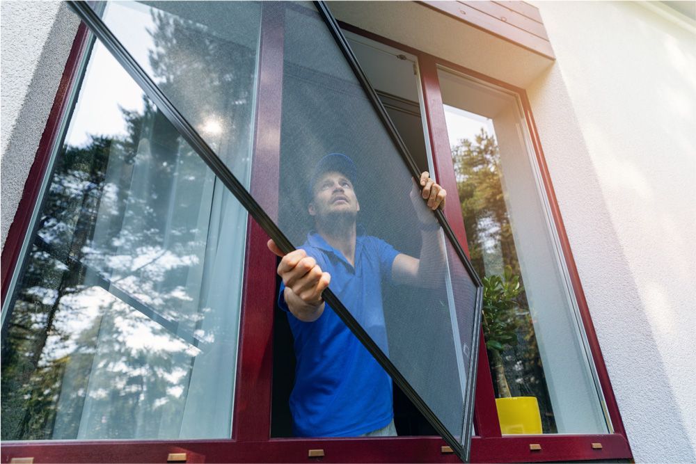 Man installing a window screen outdoors, red window frame, blue shirt, sunny.