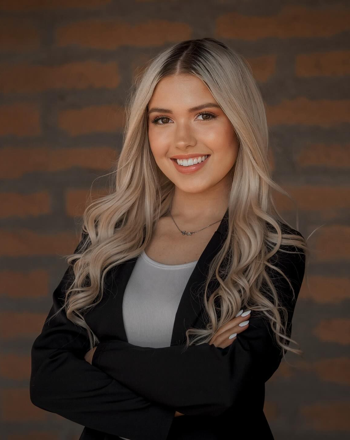 Woman with long blonde hair, smiling, arms crossed, wearing a black blazer and white shirt in front of a brick wall.