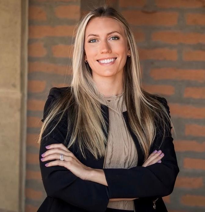 Woman with blonde hair, arms crossed, smiles confidently in front of a brick wall.