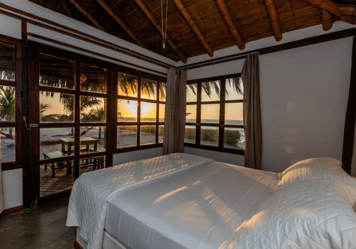 Dormitorio con cama blanca y ventanales con vistas a una playa al atardecer.