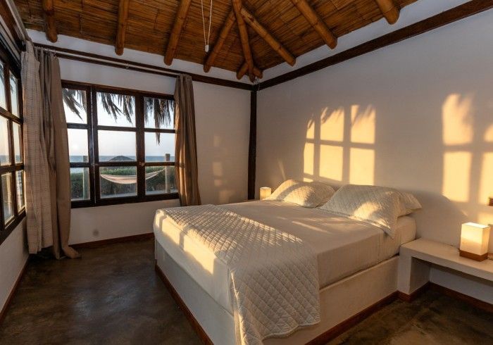 Dormitorio con cama, ventanas con vistas y techo de paja, bañado por la cálida luz del sol.