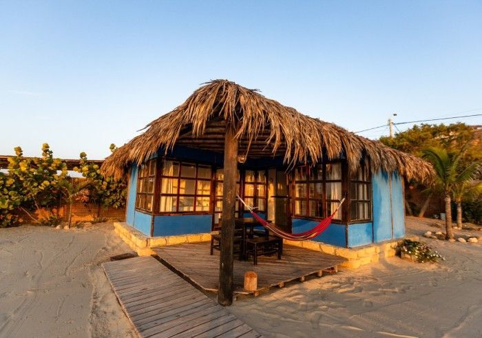 Cabaña de playa azul con techo de paja, porche y hamaca; entorno de arena.
