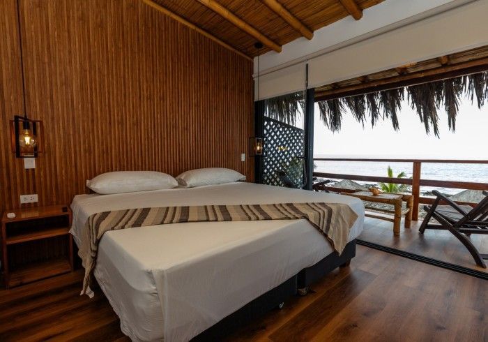 Dormitorio con vista al mar; paredes y piso de madera, cama con sábanas blancas, balcón.