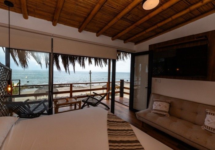 Dormitorio con vista a la playa: cama, sofá, sillas en terraza, puertas abiertas, océano visible.
