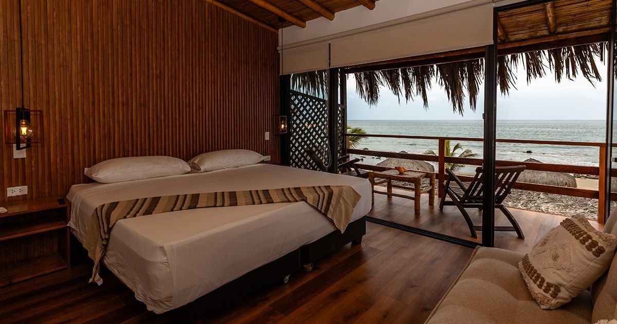 Balcón con vista a la playa; suelo de madera, sillas y mesa pequeña. Techo de paja, celosía y vista al mar.