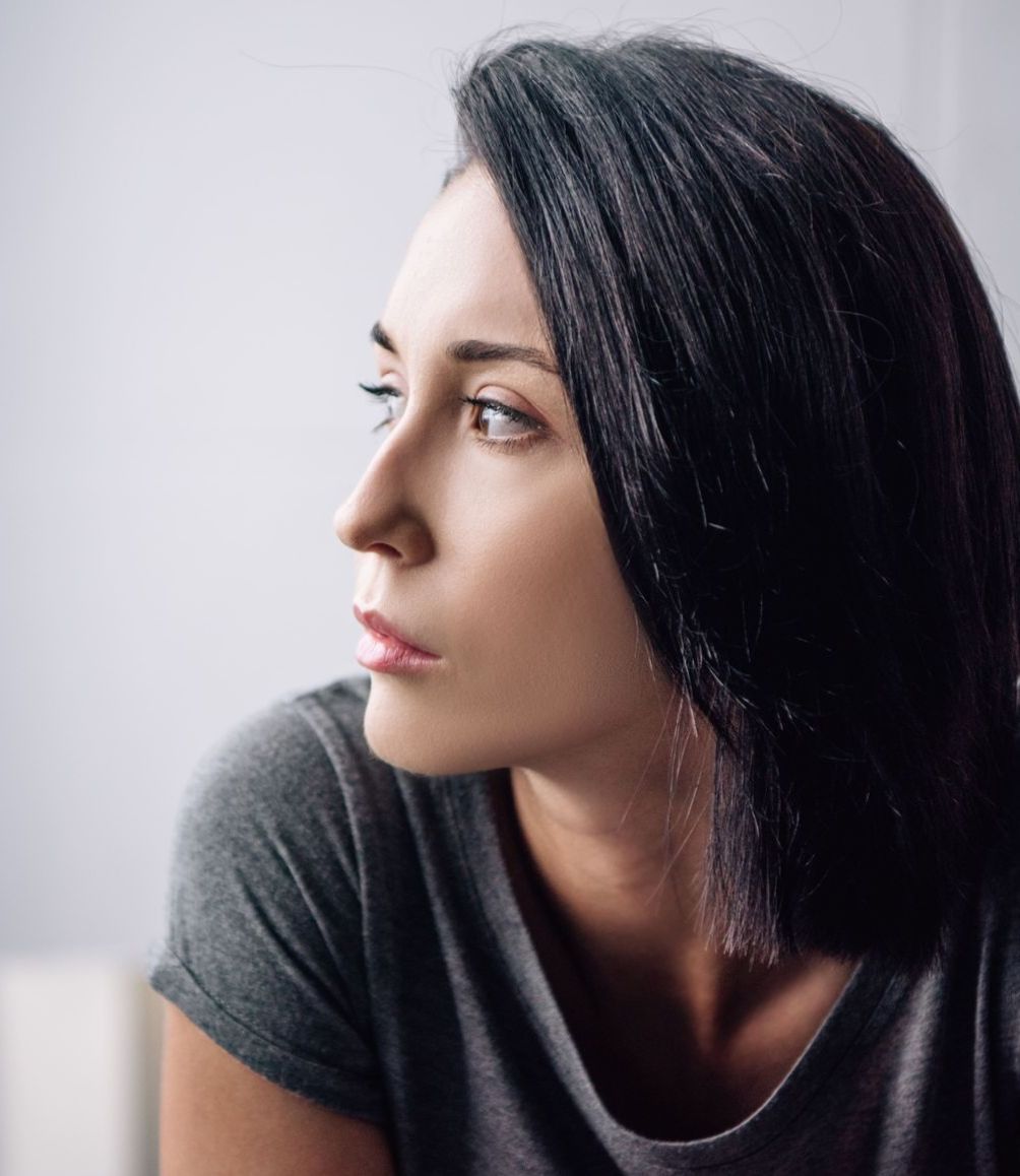 A woman with long black hair is wearing a grey shirt and looking out a window.