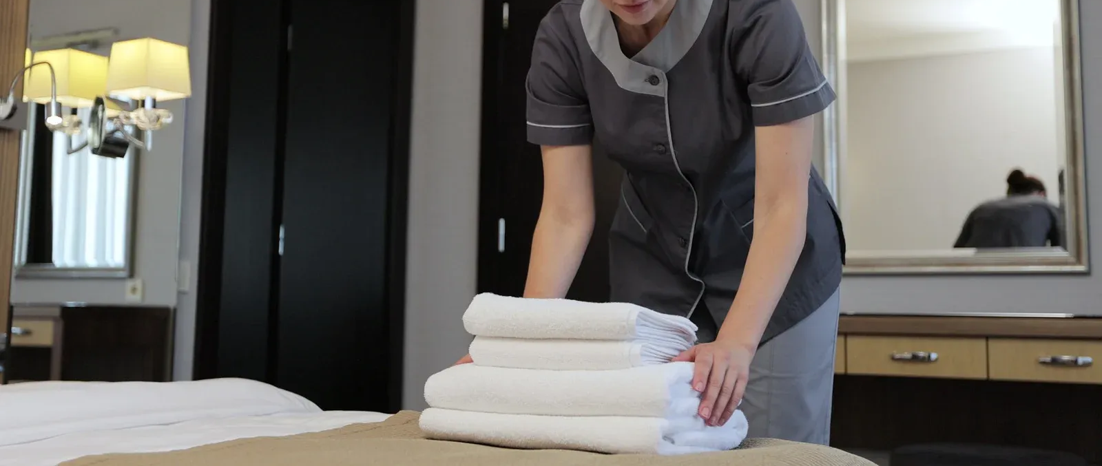 Housekeeper in gray uniform folds white towels on a bed in a hotel room.