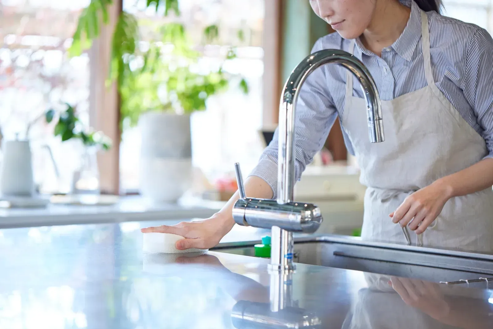 Woman in apron cleans a stainless steel kitchen countertop with a sponge.