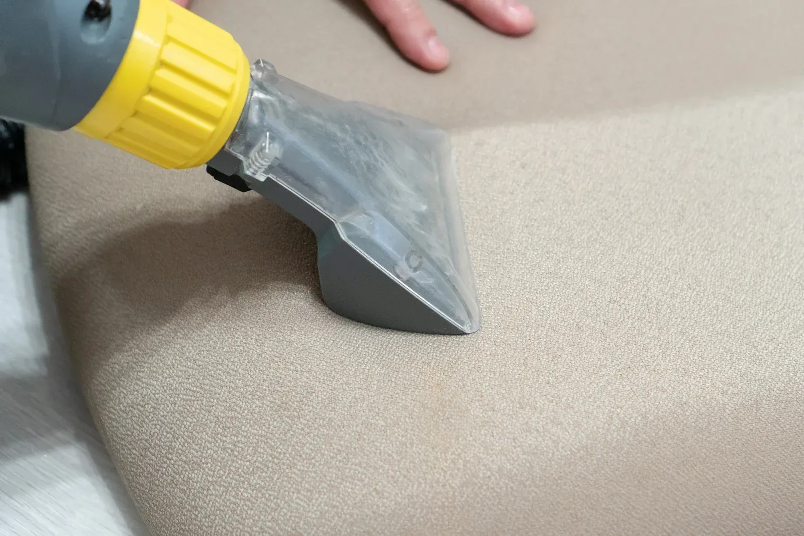 A hand using a carpet cleaner head on a light beige upholstered surface.
