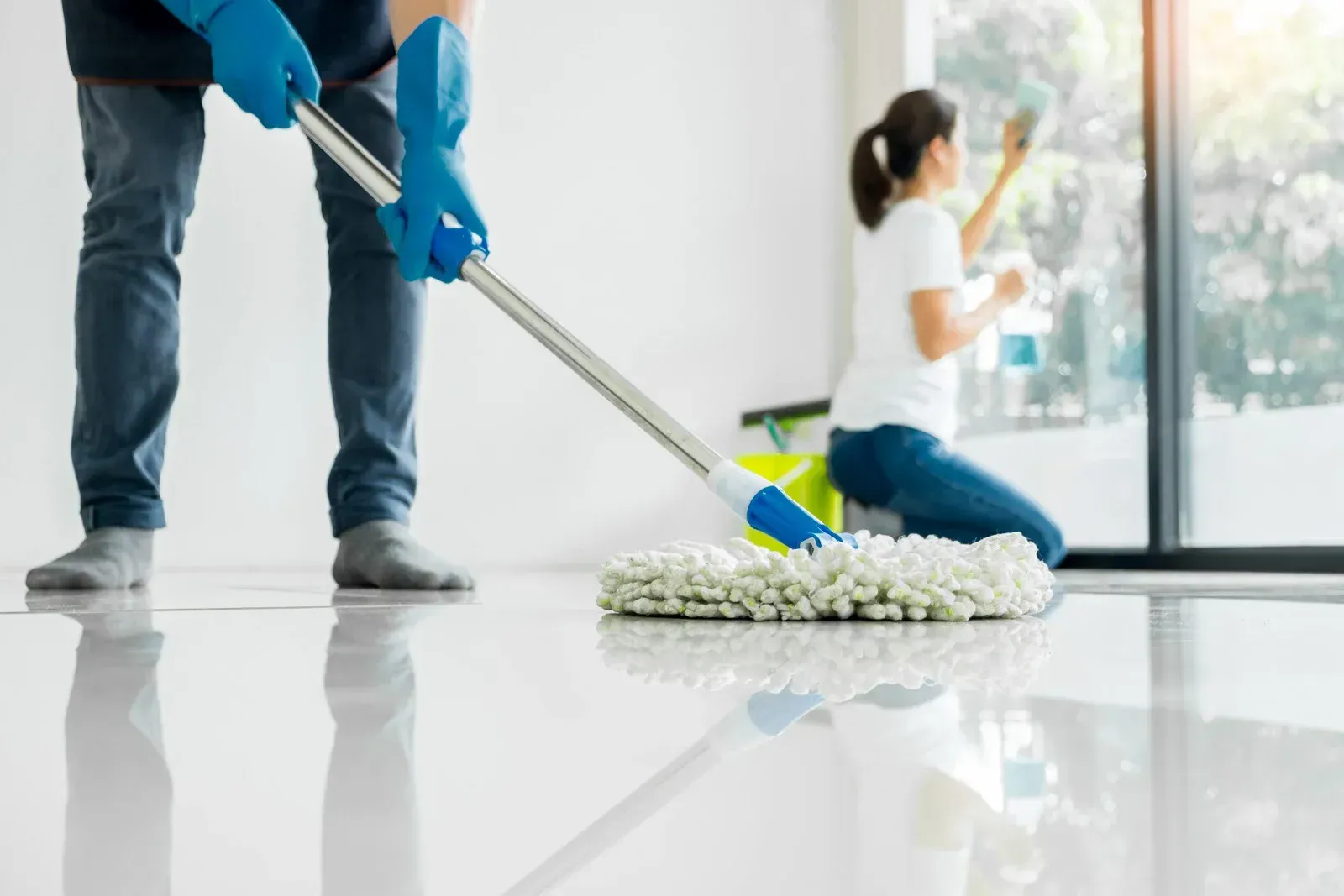 Person mopping a glossy floor, another cleaning a window. Bright room, blue gloves.
