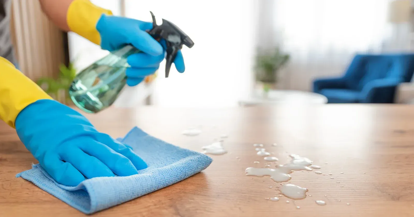 Hands in blue gloves cleaning a wooden surface with spray bottle and cloth.