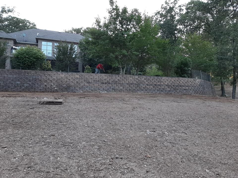 A large house is behind a stone wall in the middle of a dirt field.