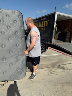 A man is carrying a mattress in front of a don katy truck.