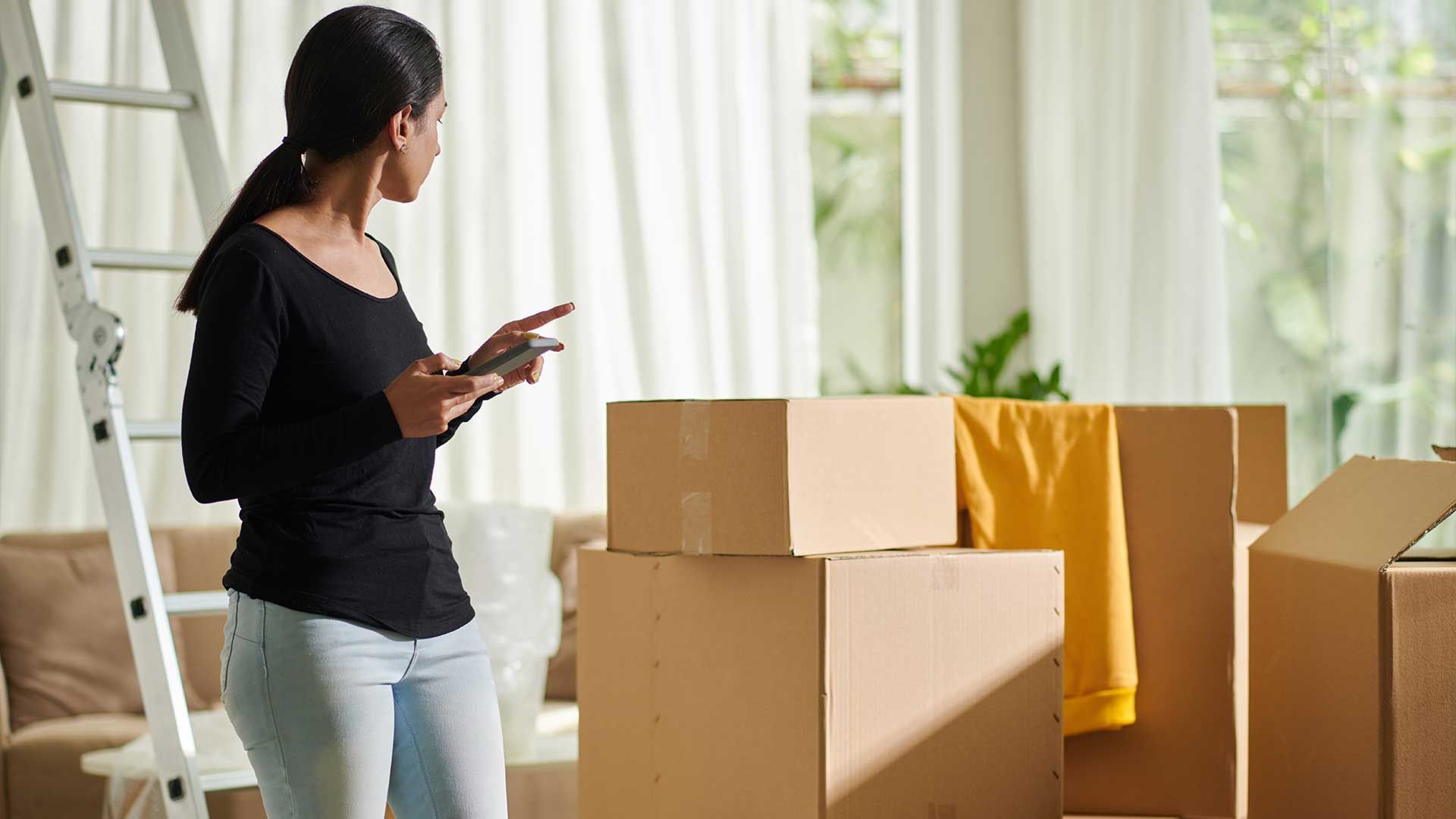 A woman is using a tablet in a living room filled with cardboard boxes.
