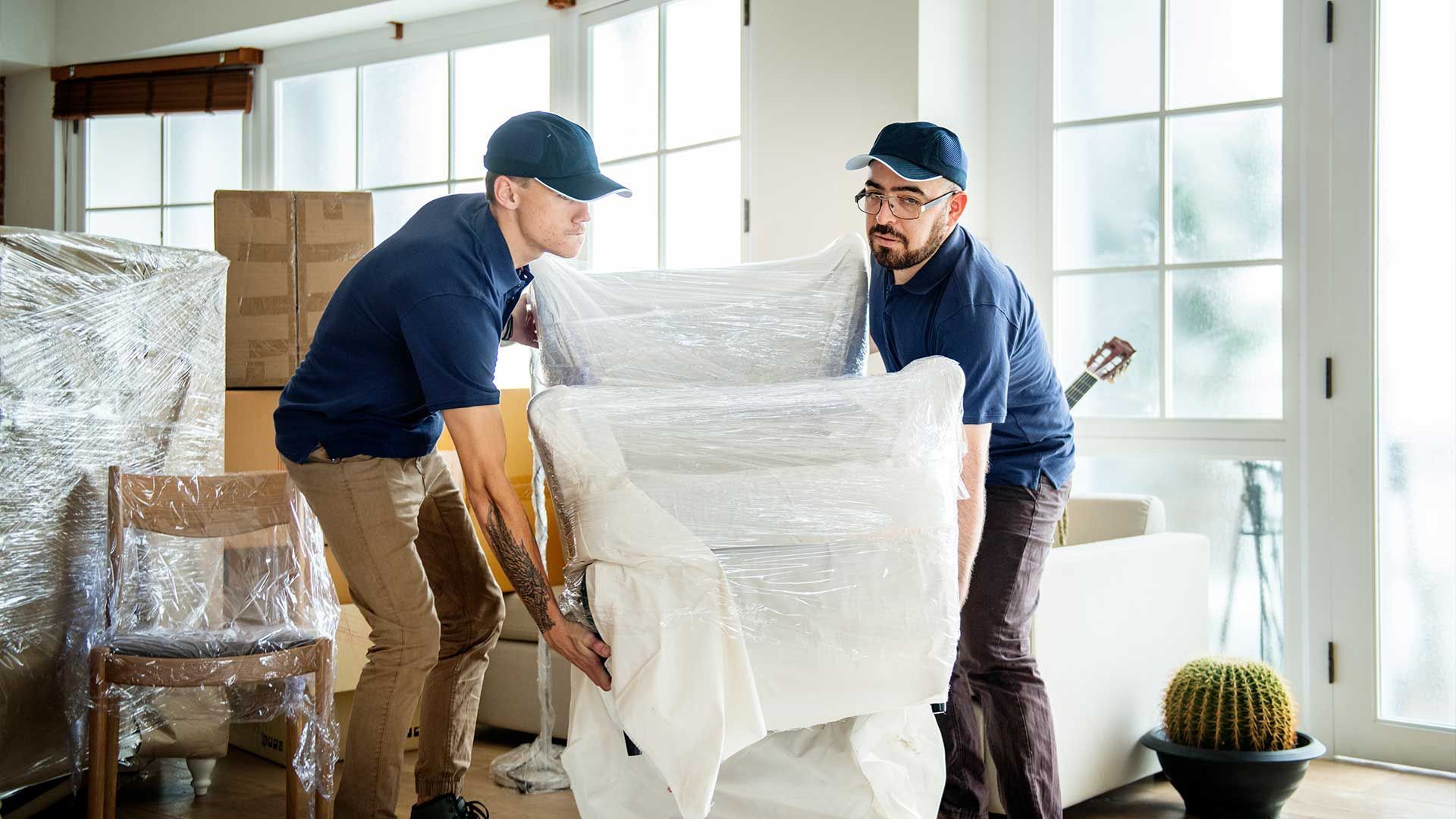 Two men are wrapping a piano in plastic wrap in a living room.