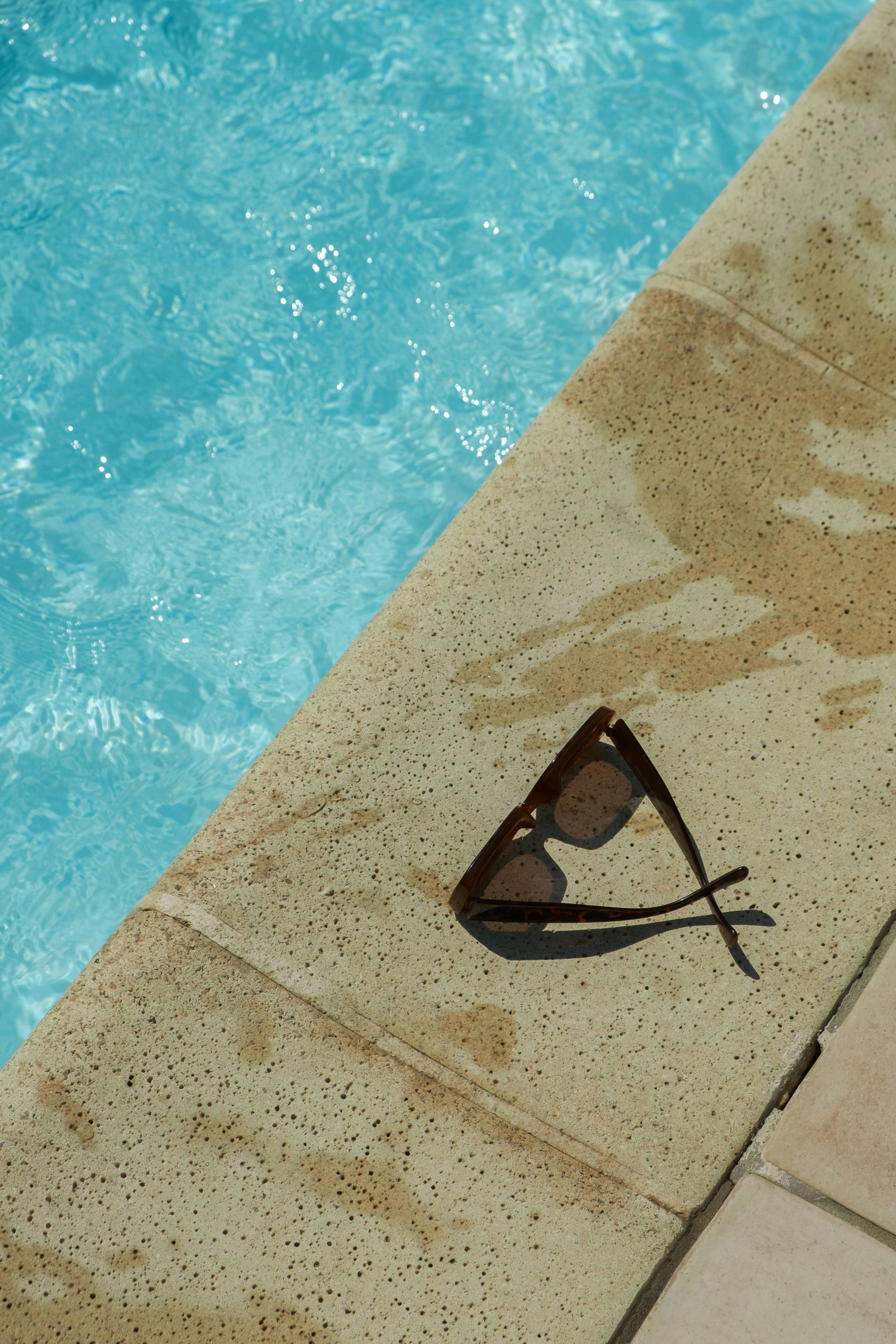 Sunglasses rest on the edge of a beige-tiled pool, water reflecting a light blue.