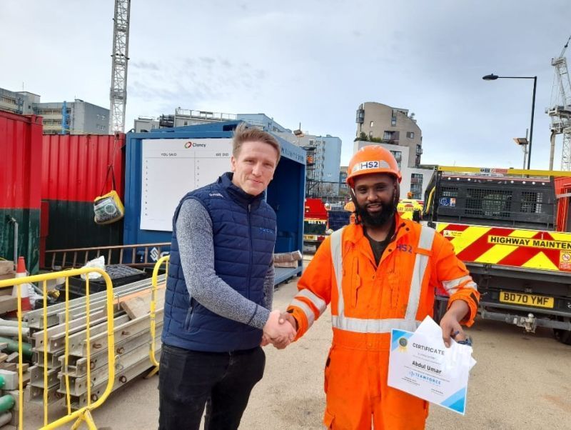 Two people in orange jumpsuits in a large industrial setting, one pointing, the other holding a blueprint.