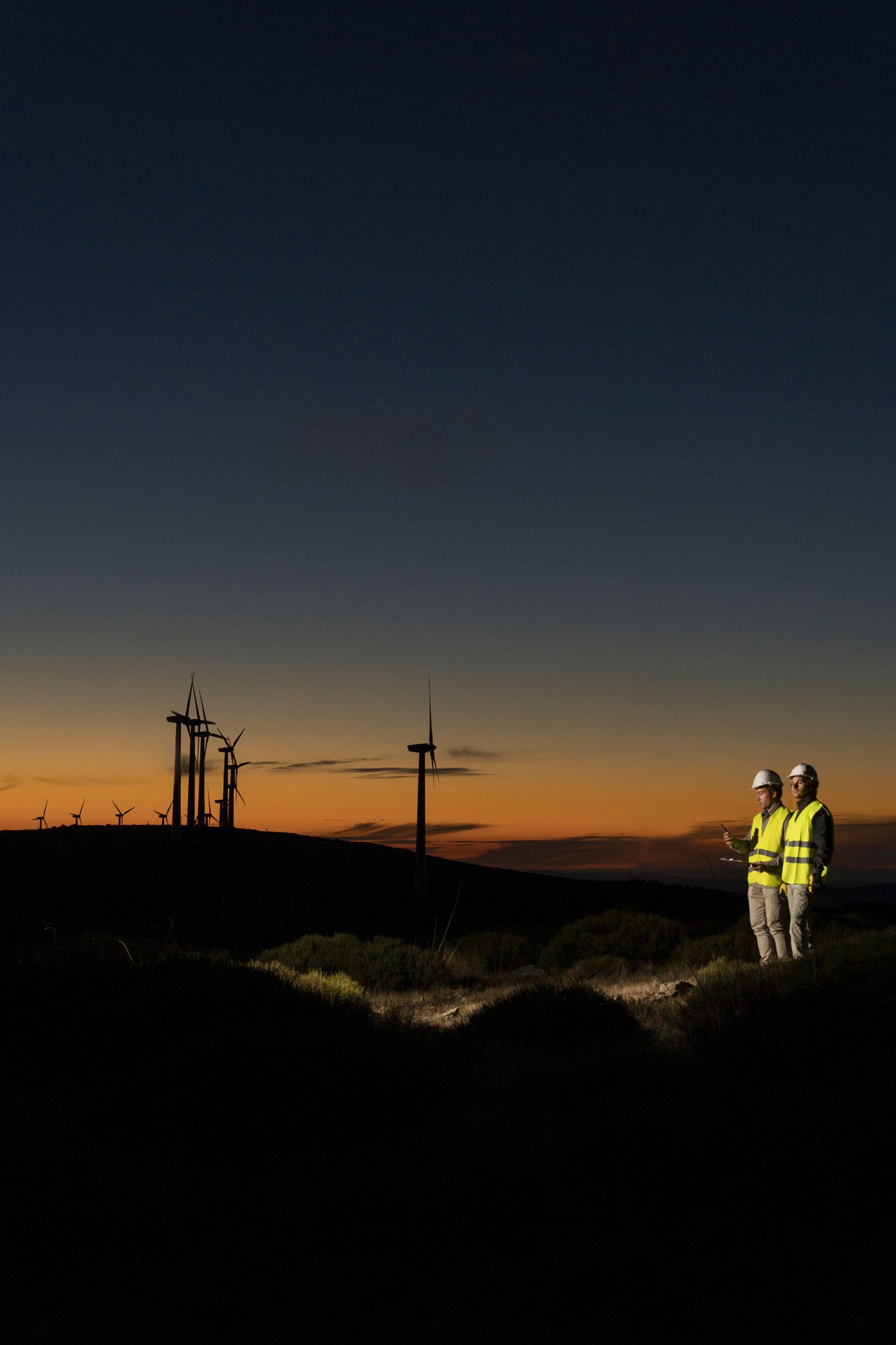 Two people in safety vests at dusk, wind turbines silhouetted on a hillside, orange and blue sky.