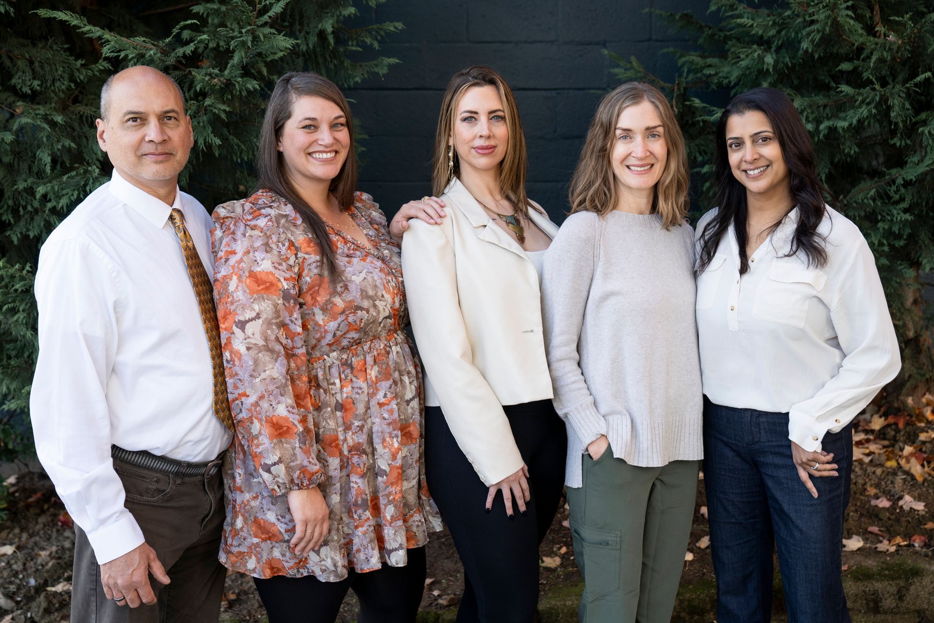 Group of five people standing outside, smiling at the camera. Trees in background.