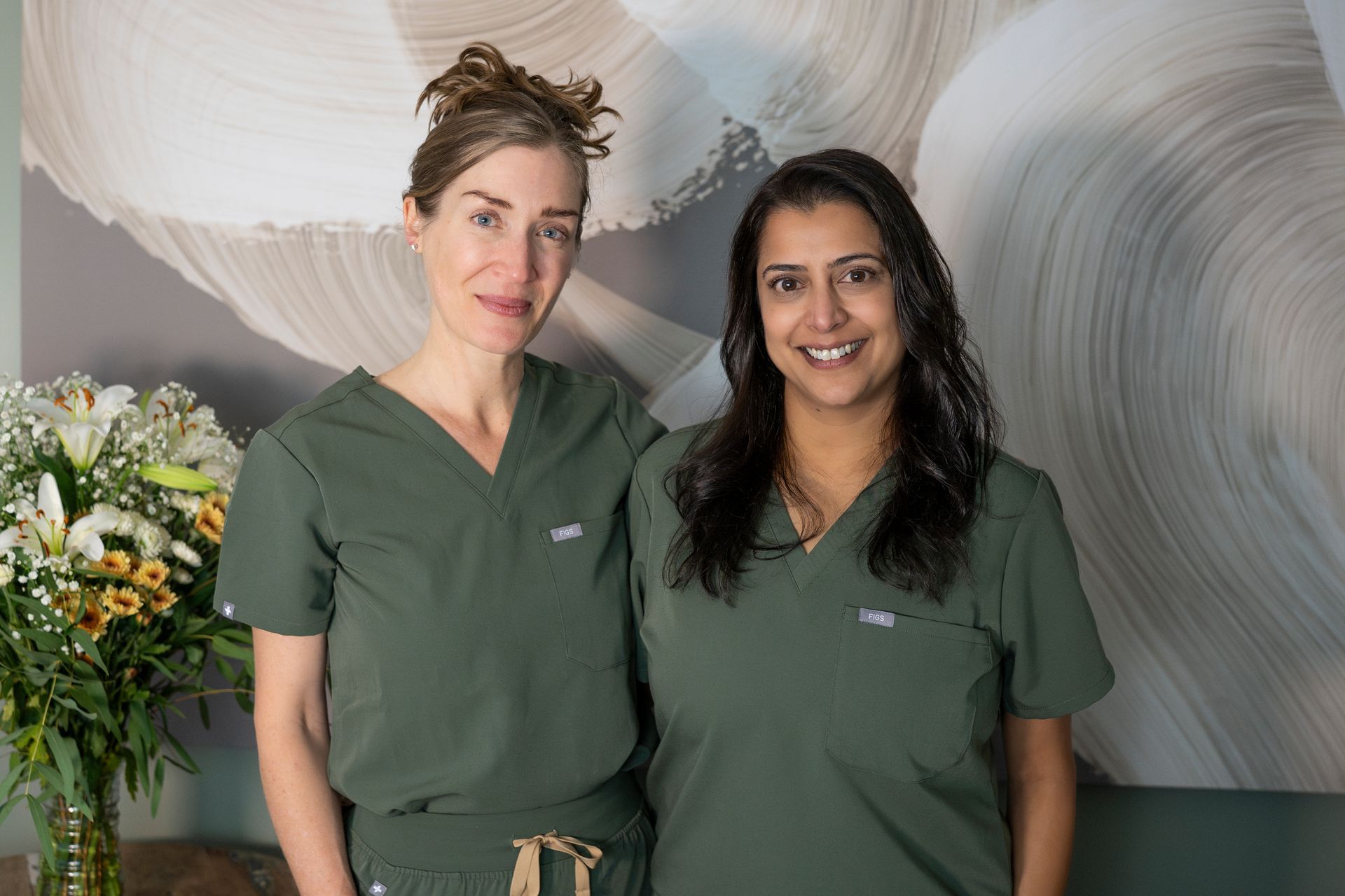 Two people in green scrubs pose indoors near flowers.
