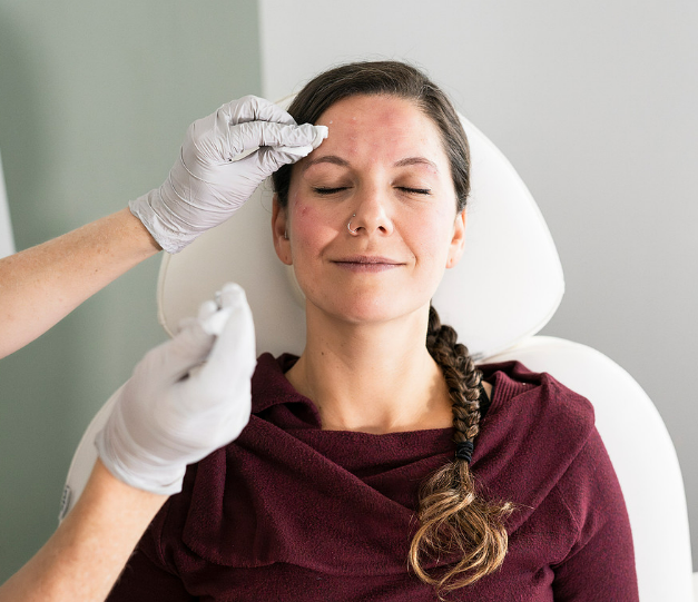 Woman receiving a facial injection in a medical setting; closed eyes, red skin, and gloved hands are visible.