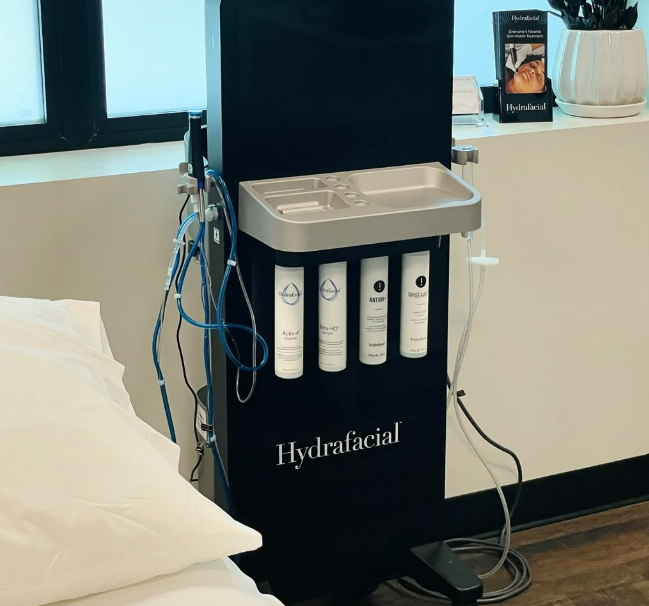 Black Hydrafacial machine with four white bottles, silver tray, and blue tubes next to a treatment bed.