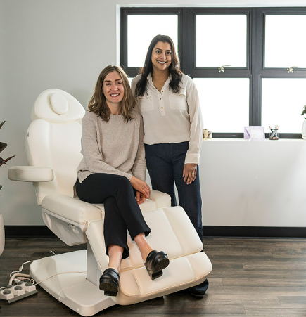 Two women in medical office setting, one seated in examination chair, smiling.